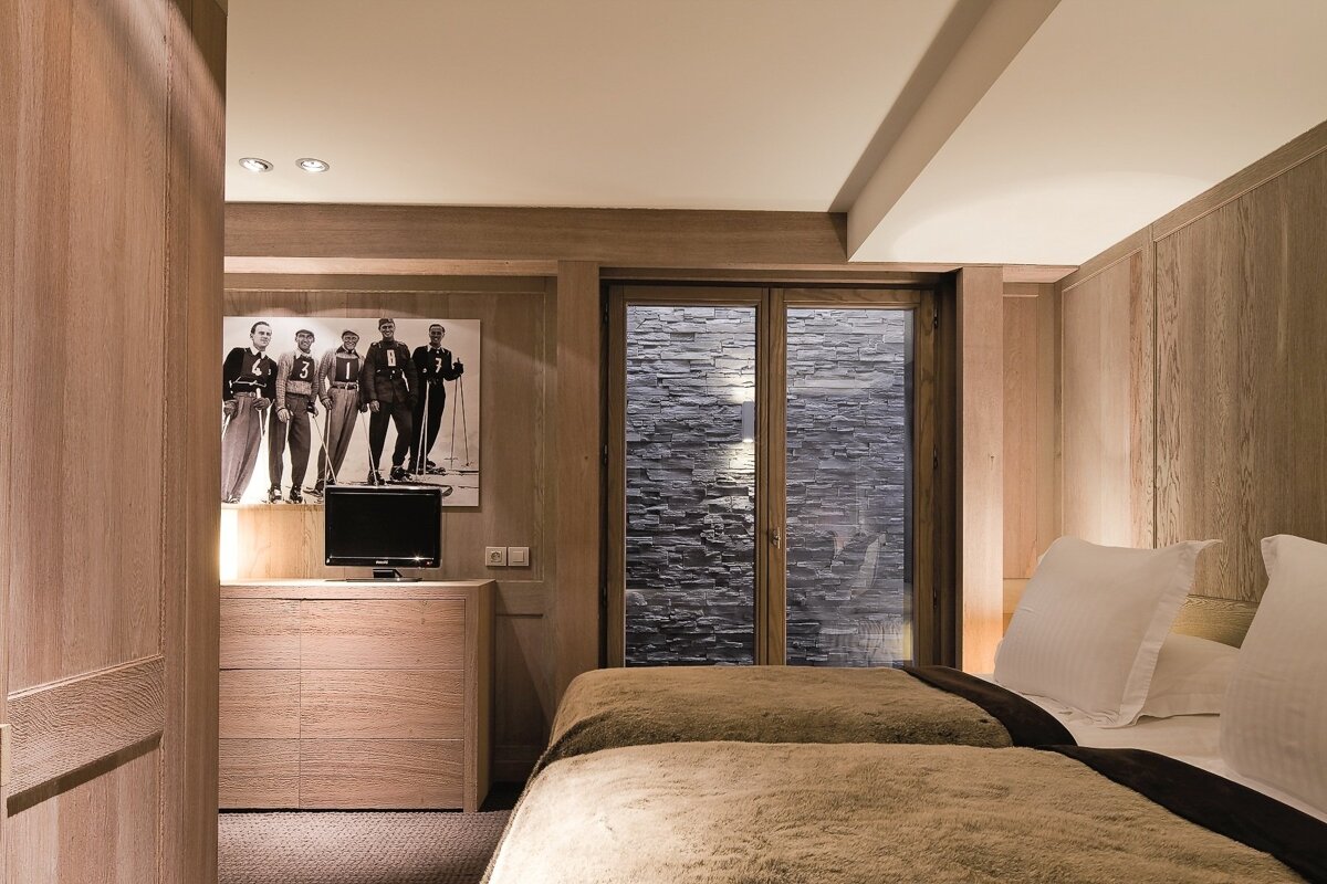 A warm, wood-paneled bedroom with twin beds, a large window facing a stone wall, a TV on a dresser, and a vintage ski poster.