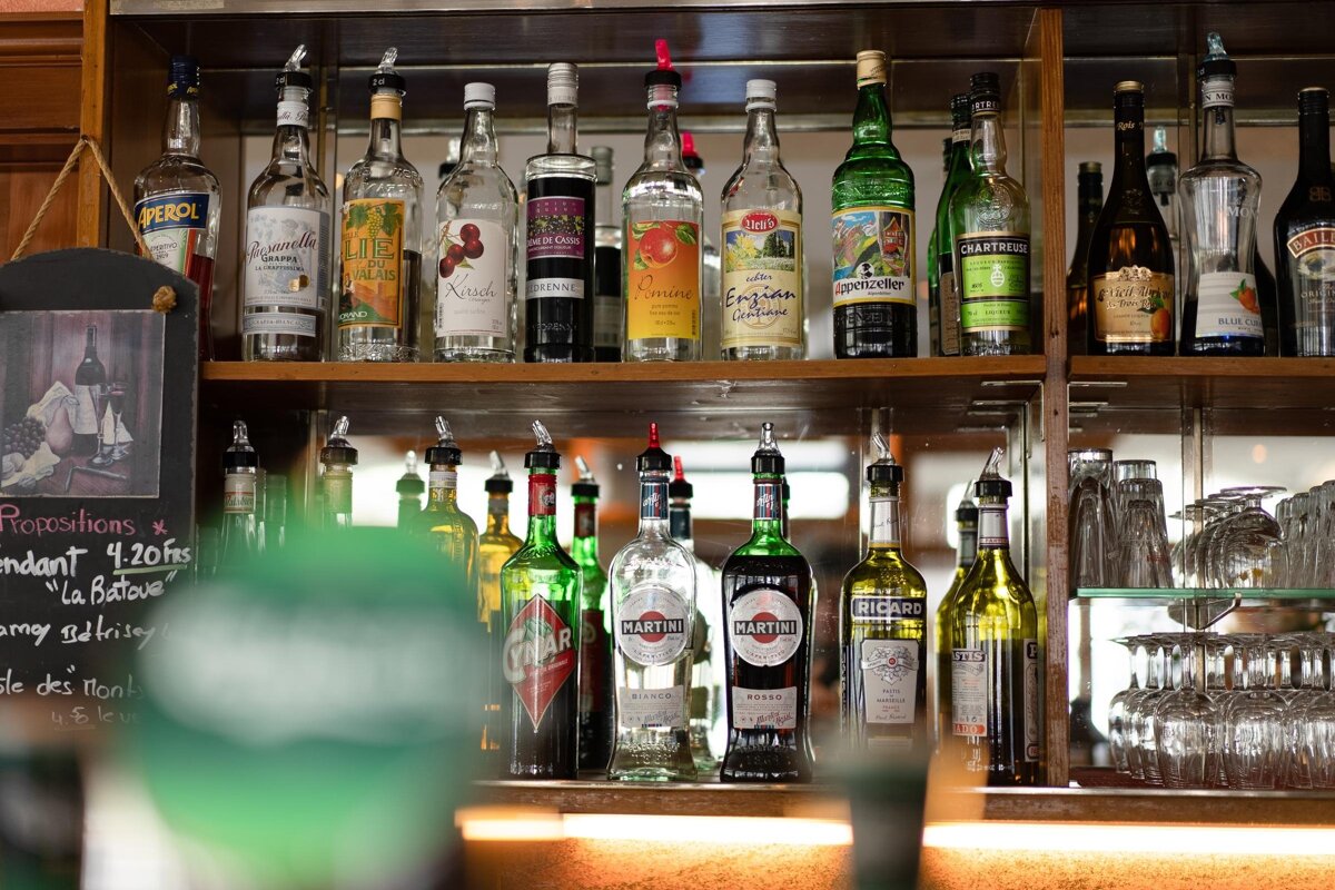Rows of liquor bottles, including Martini and Cynar, lined up on wooden bar shelves next to stacked glassware.