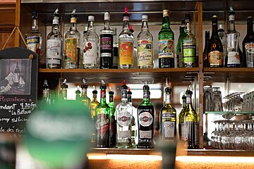 Rows of liquor bottles, including Martini and Cynar, lined up on wooden bar shelves next to stacked glassware.