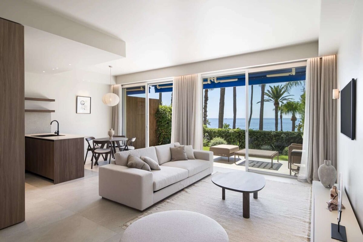 Chic, modern open-plan living room with kitchen and dining areas. Features large windows showcasing a stunning ocean view framed by palm trees.