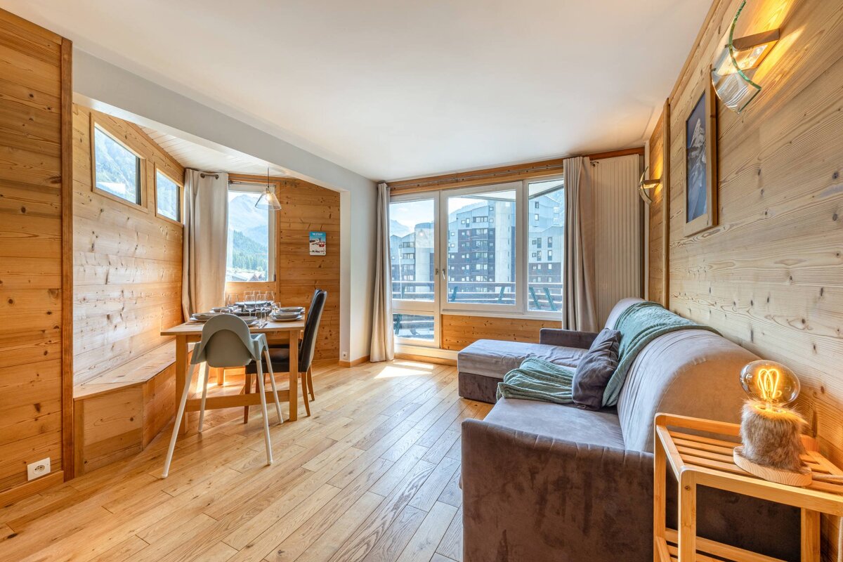 A cozy, wood-paneled living/dining room with a sofa, dining table, and large windows offering views of mountains and buildings.