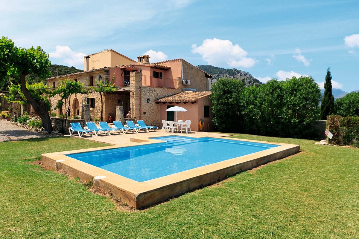 A charming stone villa with a bright blue swimming pool, sun loungers, and an outdoor dining area on a green lawn, backed by mountains under a clear sky.