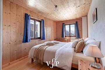 Cozy wood-paneled chalet bedroom with a neatly made double bed, blue curtains, and two windows offering scenic mountain views.