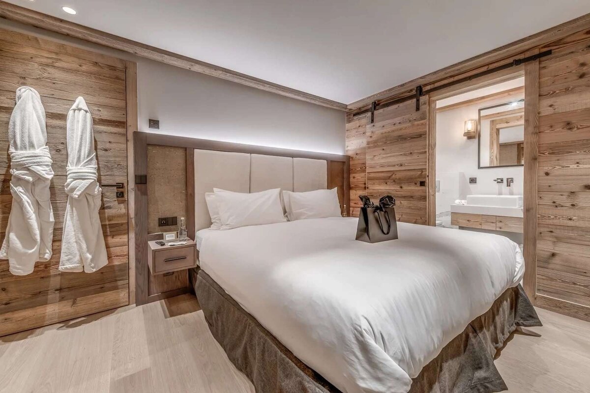 A cozy bedroom with rustic wooden walls, a large white bed, and white bathrobes. An adjoining bathroom is visible through a sliding barn door.