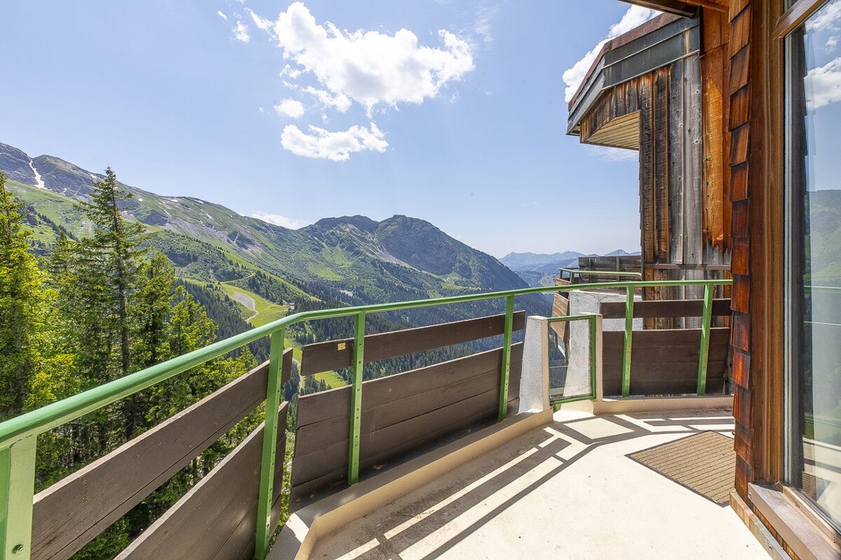 A sunny mountain view from a balcony with a green railing. Lush green slopes and trees extend under a clear blue sky, with a rustic wooden building on the right.
