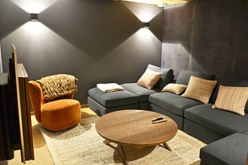 A cozy modern living room with dark walls, a gray sectional sofa, an orange armchair, a round wooden coffee table, and warm wall lights.