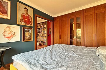 A bedroom with teal walls, paintings, a bed, wooden wardrobes, and an open doorway to a library with red walls and many bookshelves.