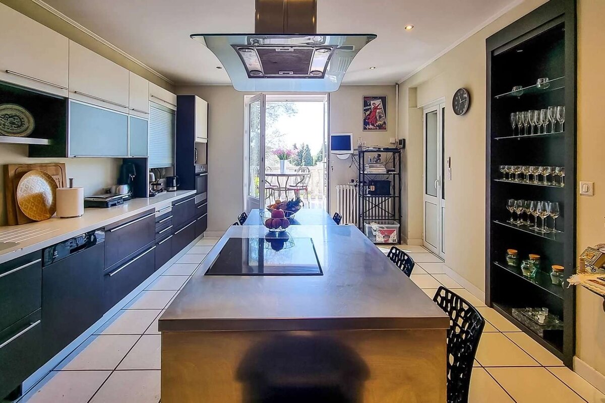 A modern kitchen with a long island cooktop, white & dark cabinets, and an open balcony door. A display shelf with glassware is on the right.
