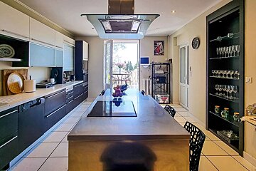 A modern kitchen with a long island cooktop, white & dark cabinets, and an open balcony door. A display shelf with glassware is on the right.