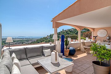Sunny terrace with a gray L-shaped sofa, dining area, unique decor including cacti, and a breathtaking view of the city and ocean.