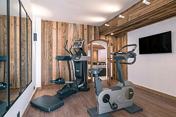 A rustic yet modern home gym featuring an elliptical, stationary bike, wall-mounted TV, and large mirror, with wooden walls and ceiling.