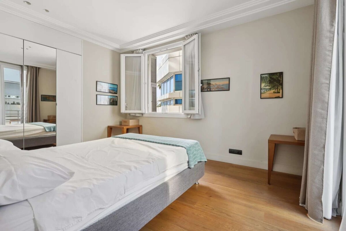 A bright, modern bedroom features a bed with white linen, hardwood floors, an open window showing buildings, a mirrored closet, and wall art.