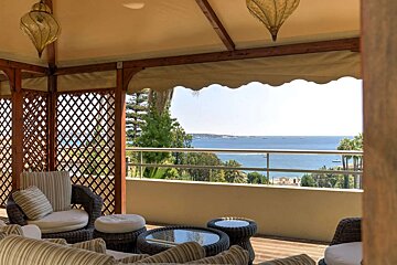 A shaded outdoor terrace with comfy wicker furniture, lattice walls, and lanterns, overlooking a stunning blue ocean dotted with boats.