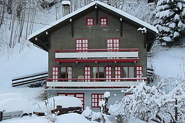 A gray house with red-trimmed windows and balconies is covered in thick snow, nestled in a snowy mountain landscape with snow-covered cars and trees.