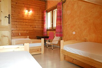 A warm, rustic dorm room featuring wooden walls, an orange textured wall, several simple wooden beds, a window with pink curtains, and chairs.