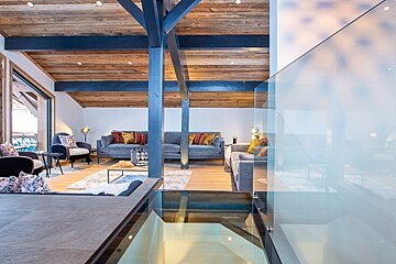 Modern chalet loft living room featuring wooden ceilings, dark beams, cozy gray sofas, and a unique glass floor panel.
