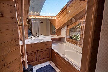 Cozy wooden bathroom with a bright, slanted skylight. Features a sink, lighted mirror, and a tub with a glass partition and small window.
