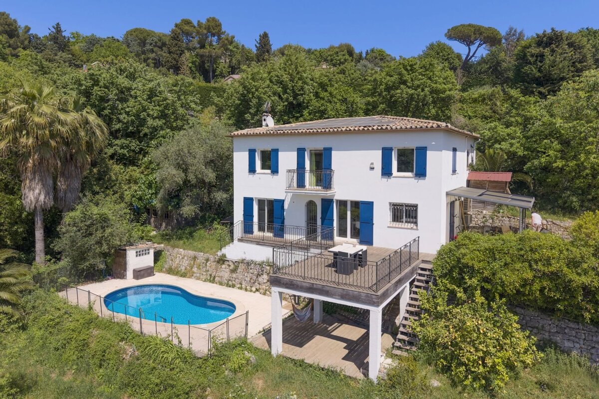 A two-story white villa with blue shutters, a terrace, and a swimming pool, surrounded by lush green trees under a clear blue sky.