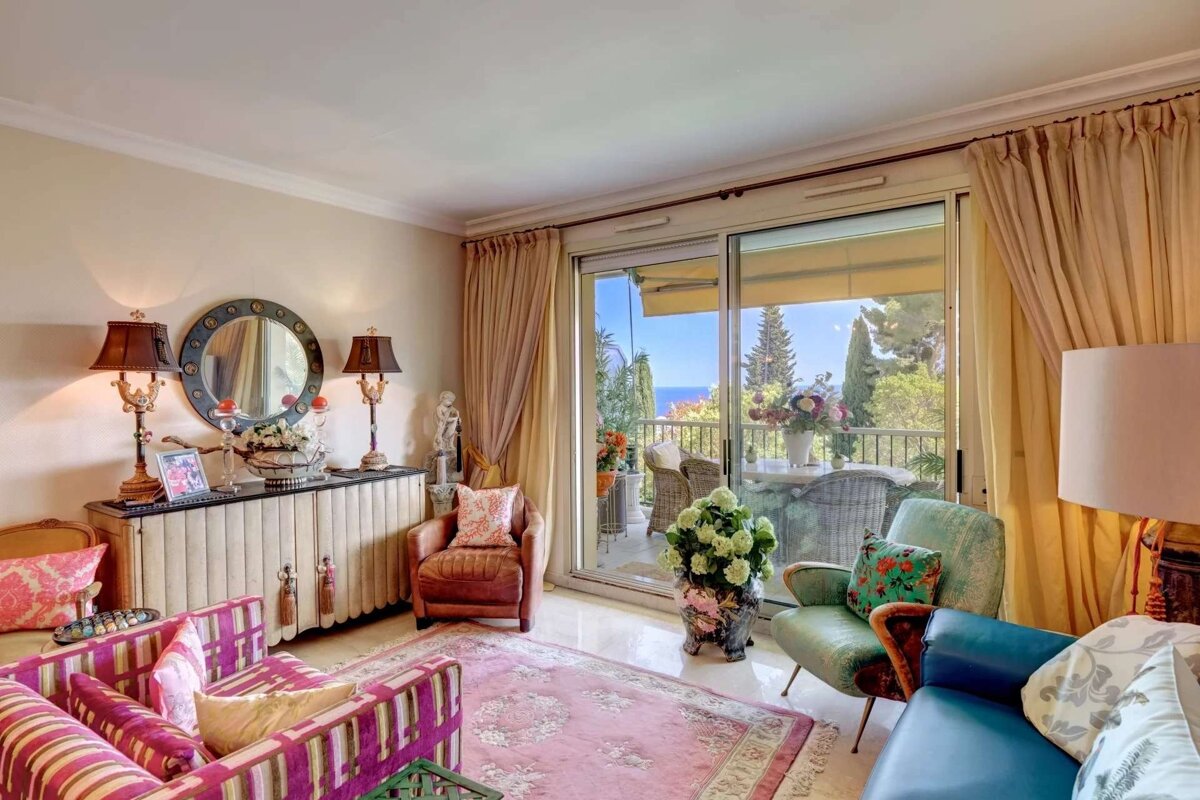 Vibrant, eclectic living room with varied furniture & patterns, opening to a sunny balcony overlooking the Mediterranean Sea & lush gardens.