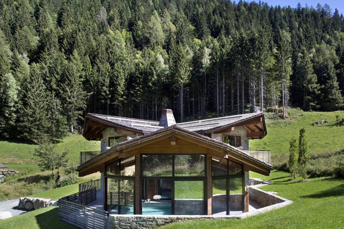 Mountain chalet with a glass-enclosed pool on a green hill, surrounded by a dense forest.