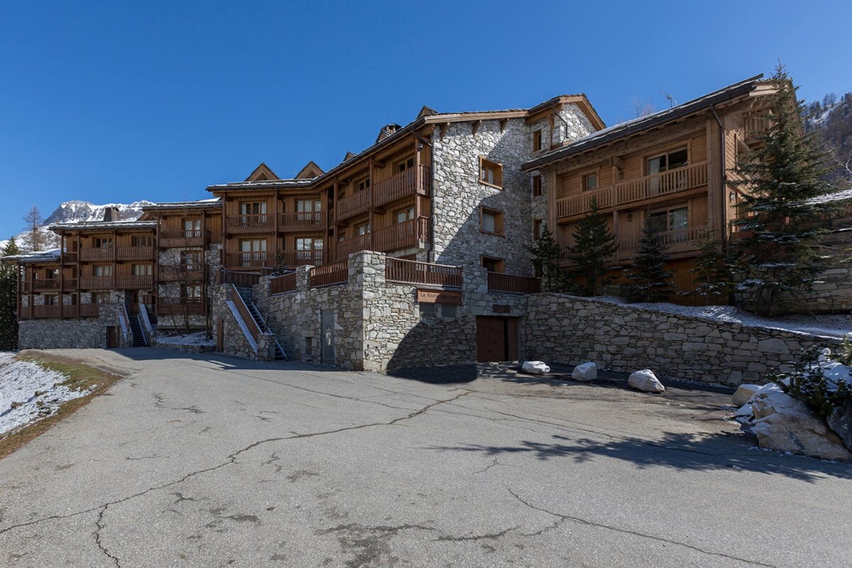 A large, traditional stone and wood chalet complex with balconies, nestled in a snowy mountain landscape under a clear blue sky.
