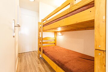 A narrow room features light wooden bunk beds with brown bedding. The space has light walls, wood-look flooring, and a closed white door.