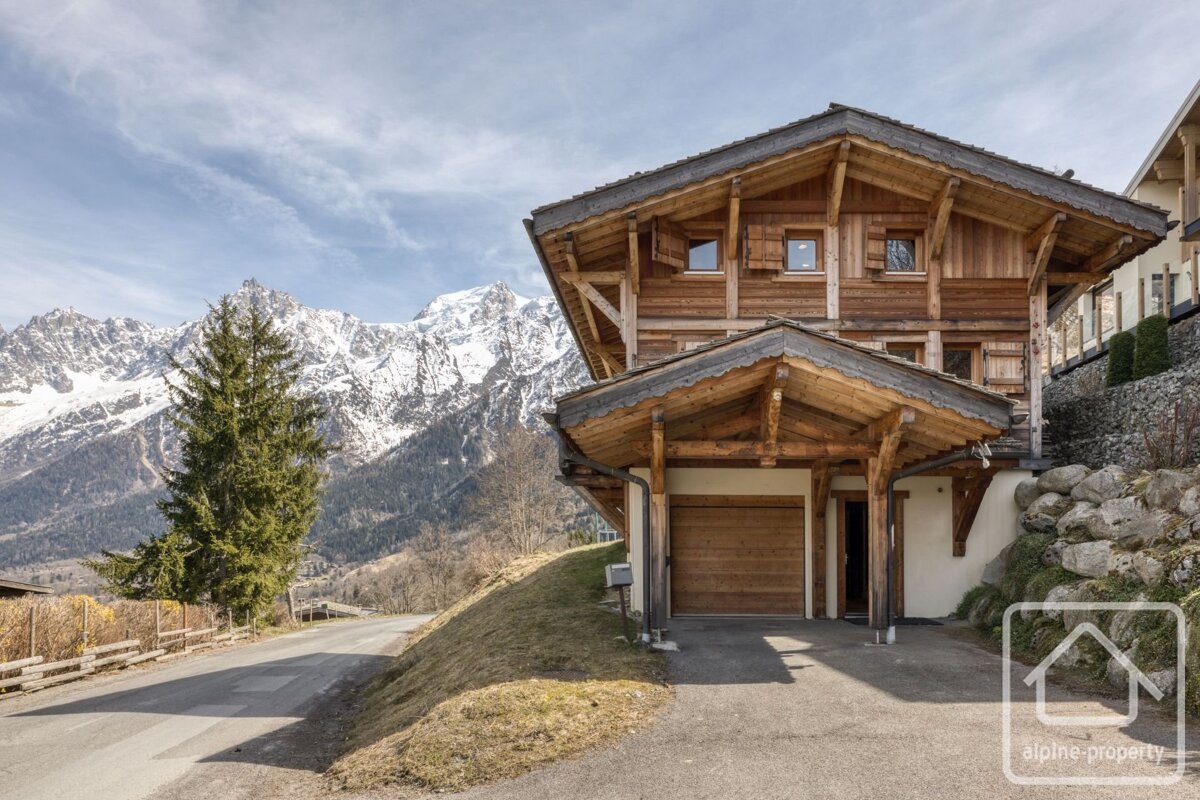 A large wooden chalet with a garage next to a road, with snow-covered mountains in the background.