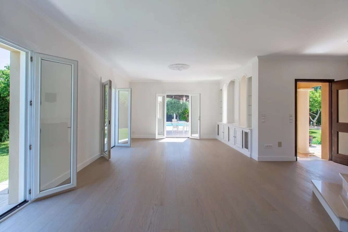Spacious, bright room with light wooden floors, white walls, and built-in cabinetry. Open doors connect to a green garden and pool area.