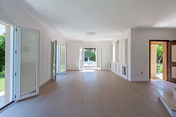 Spacious, empty room with light wood floors, white walls, and multiple open doors offering views of a lush green outdoor area and a distant pool.
