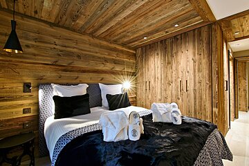 Chalet-style bedroom with rich wooden walls, ceiling, and integrated wardrobes. The bed has white and black linens, along with hotel robes and slippers.