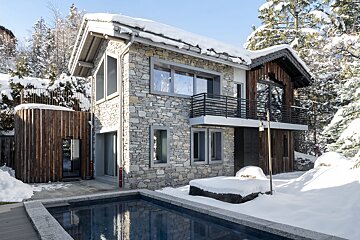 A modern stone and wood chalet in a snowy landscape with an outdoor pool, surrounded by winter trees under a clear sky.