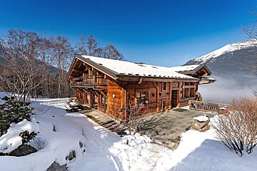 A rustic wooden chalet covered in snow, set against majestic mountains and a clear blue sky.