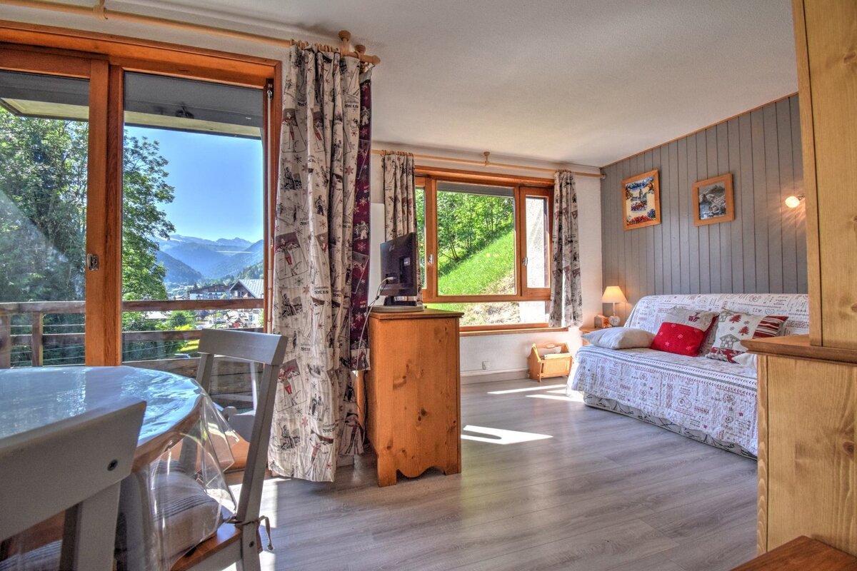 Sunny alpine apartment interior with a sofa bed, wooden furniture, and large windows overlooking scenic mountain views.