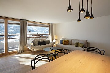 A modern, sunlit living area featuring wooden floors, stylish furniture, and large windows with a scenic snowy mountain view.