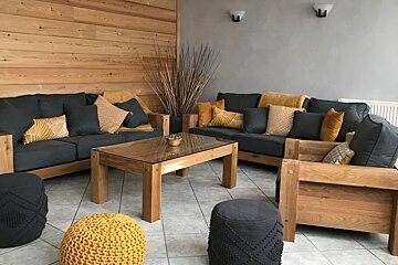 Cozy lounge featuring wooden sofas with grey cushions, yellow accent pillows, knitted poufs, and a wood-paneled wall.