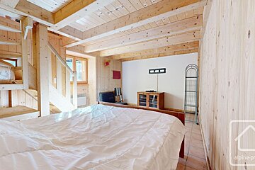 Cozy bedroom featuring light wood paneling, exposed ceiling beams, a bed with white bedding, and a wooden staircase. A window and TV mount are visible.