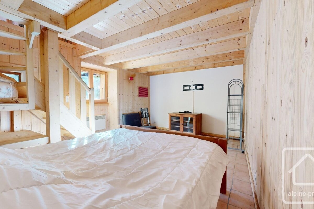 A rustic, wood-paneled bedroom features a large bed, exposed ceiling beams, and a wooden staircase. A window and entertainment area are visible.
