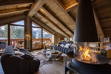 Cozy chalet living room featuring a modern central fireplace, wood-beamed ceiling, plush sofas, and a scenic snowy mountain view.