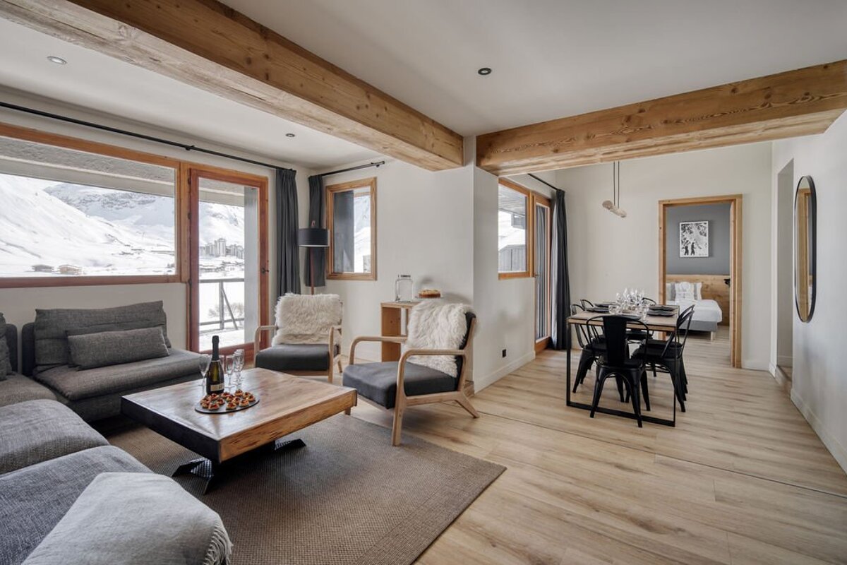 Modern, cozy ski chalet living room with mountain views, an L-shaped sofa, armchairs, and a dining area, featuring wooden beams and light floors.