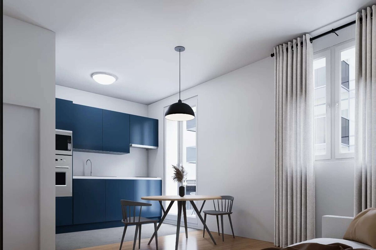 A modern, minimalist open-plan kitchen and dining area featuring blue cabinets, a wooden table with two chairs, and a large window with light curtains.