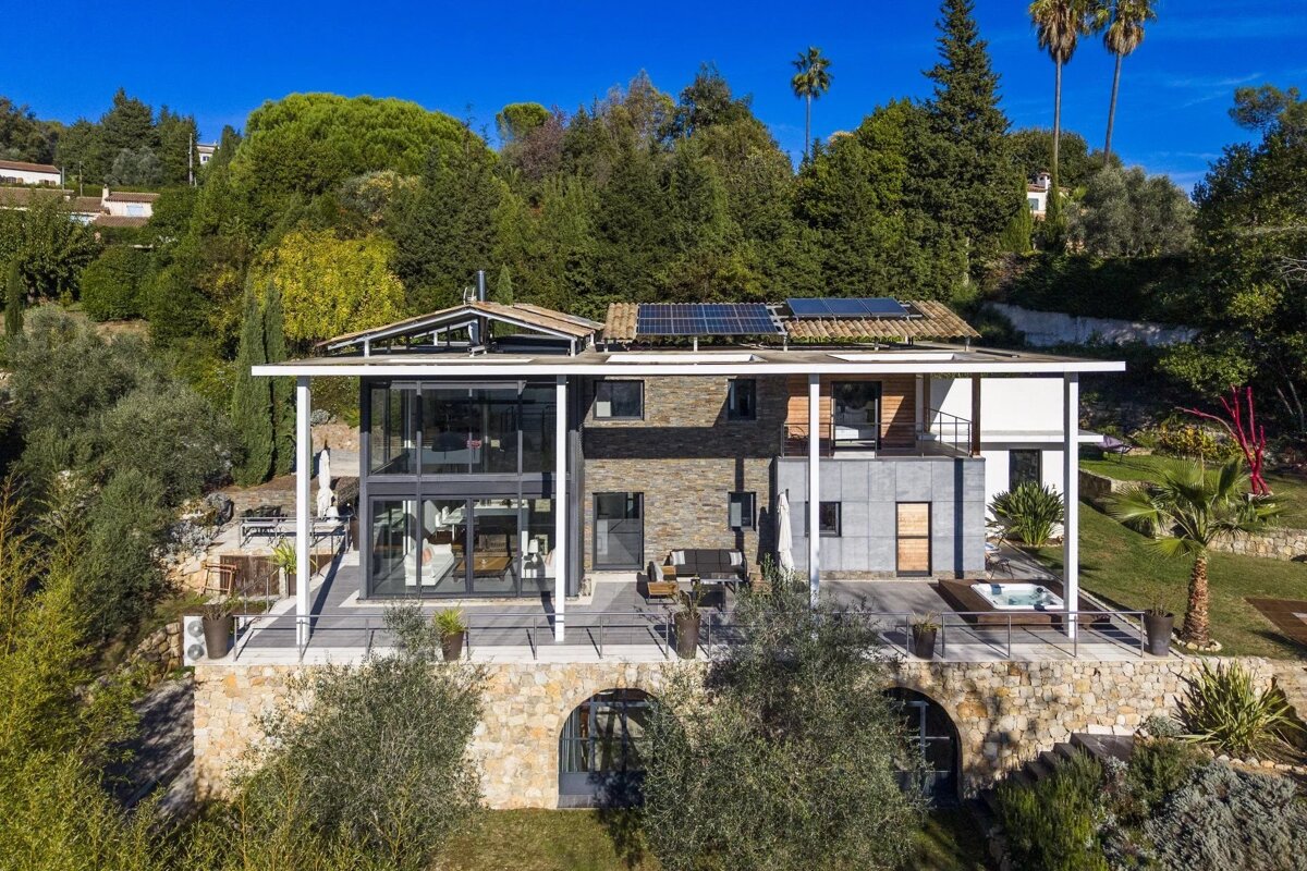 Modern glass & stone home with solar panels, terraces, and a jacuzzi, nestled on a lush, green hillside under a clear blue sky.