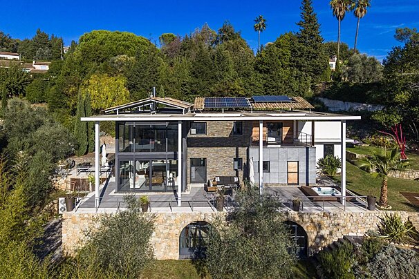 A modern, multi-level hillside villa with a large patio, glass walls, solar panels, and a hot tub, surrounded by lush green trees.