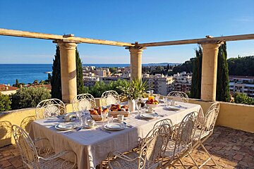 Sunlit terrace with a set breakfast table, offering panoramic views of a coastal town, the sparkling blue sea, and a clear sky.