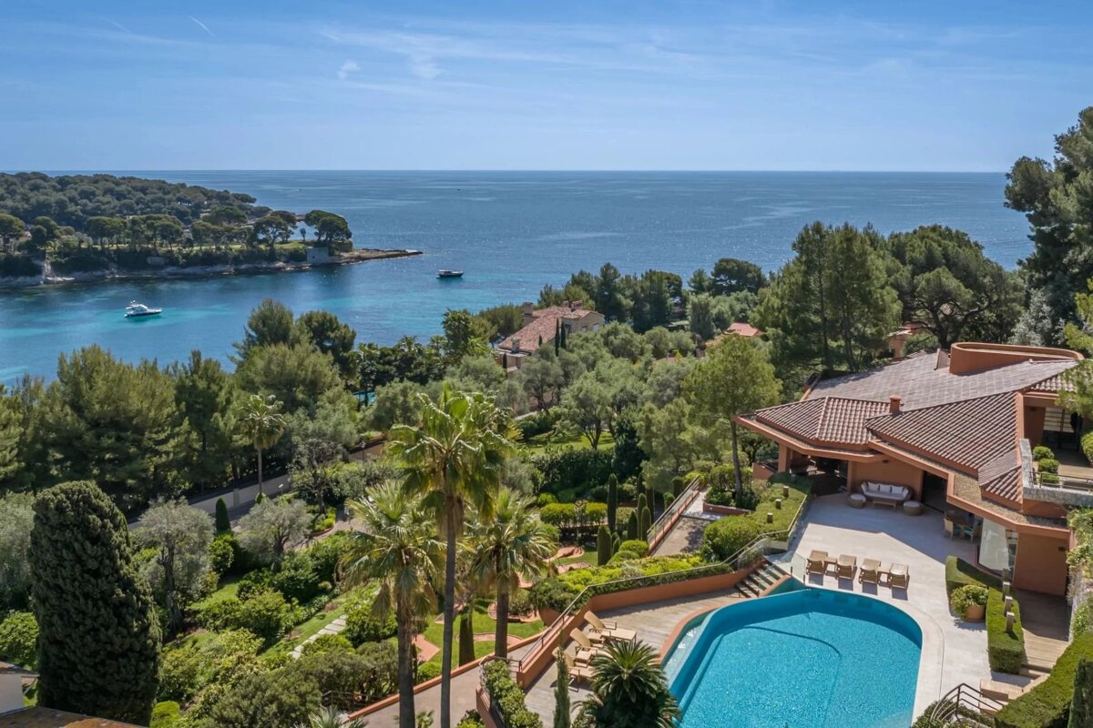 A luxury coastal villa with a swimming pool overlooking a beautiful blue sea bay surrounded by lush green trees.