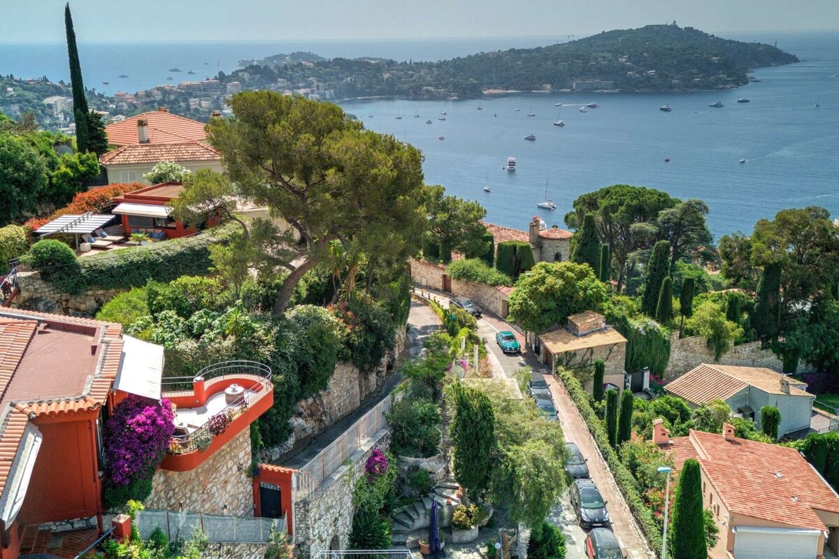 A vibrant Mediterranean village cascades down a hillside to the azure sea, featuring red-roofed houses, lush greenery, winding roads, and numerous boats.