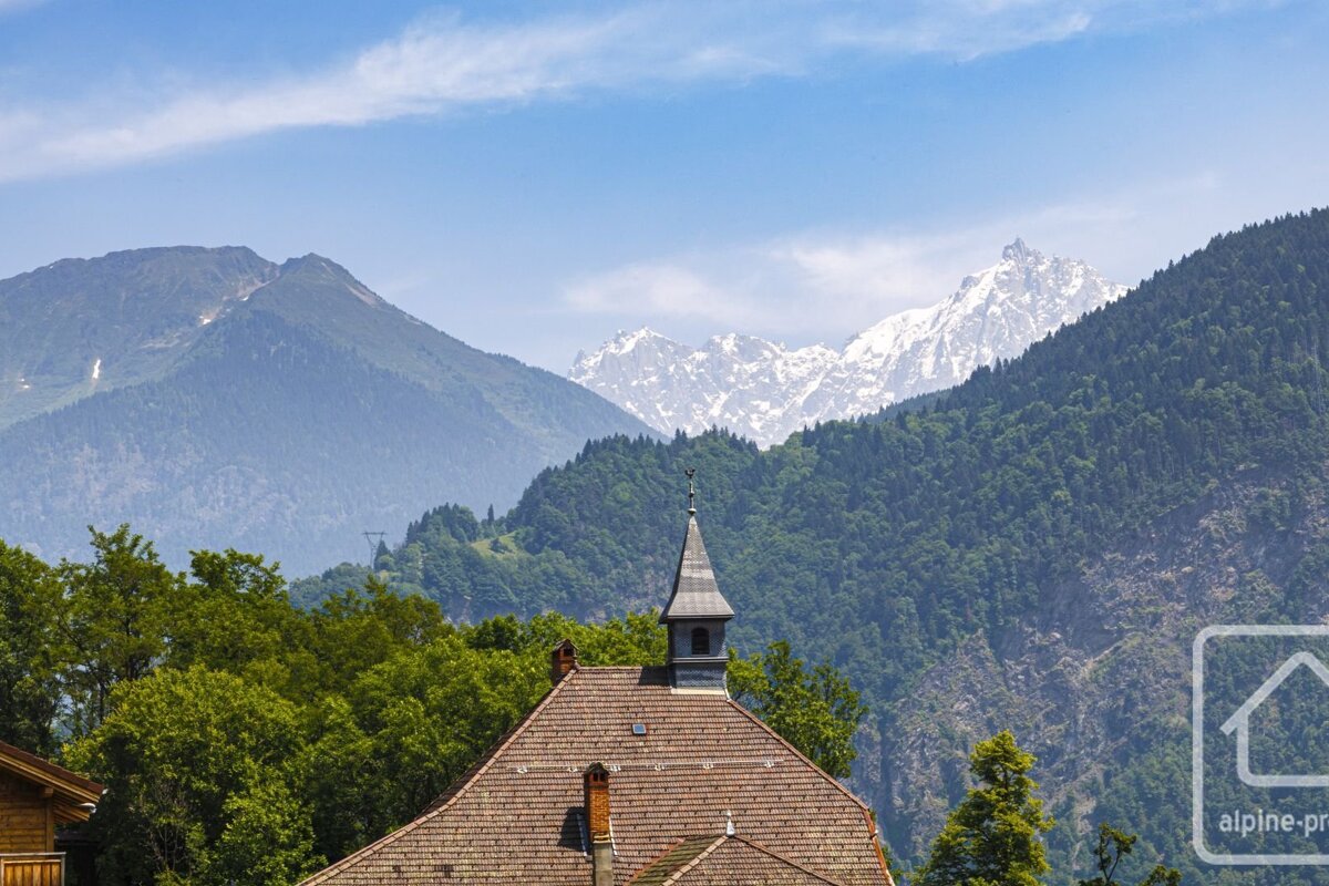 A building with a steeple and tiled roof in front of green trees, layered mountains, and distant snow-capped peaks under a clear blue sky.