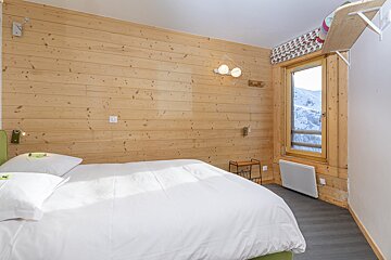 A cozy bedroom with light wood paneling, a white bed, and a window offering a snowy mountain view. Modern lighting and decor complete the look.