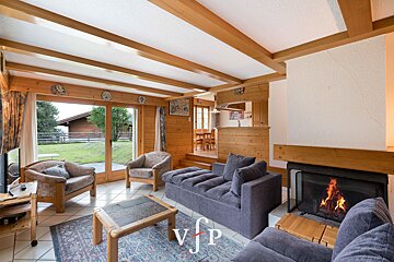 Cozy chalet living room with wooden ceiling beams, a warm fireplace, comfortable seating, and large glass doors opening to a grassy yard.