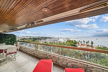 A balcony with seating and red lounge chairs overlooks a bustling marina, the sea, a coastal town, and palm trees under a blue sky.