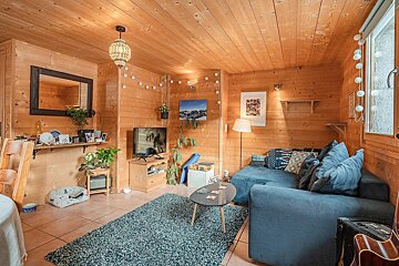 A warm, wood-paneled living room with a blue L-shaped sofa, TV, plants, and a dark rug. Eclectic decor and string lights create a cozy, rustic feel.